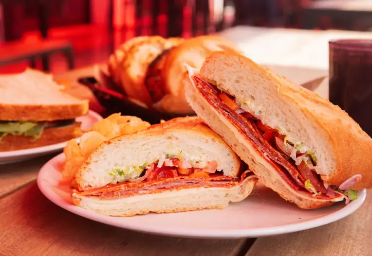 Italian sandwich cut in half with salami, lettuce, tomato, and onion on a white plate with chips.
