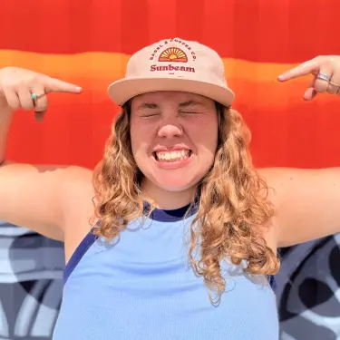 Woman smiling with eyes closed pointing at her beige Fuoco brand hat standing against a red wall.